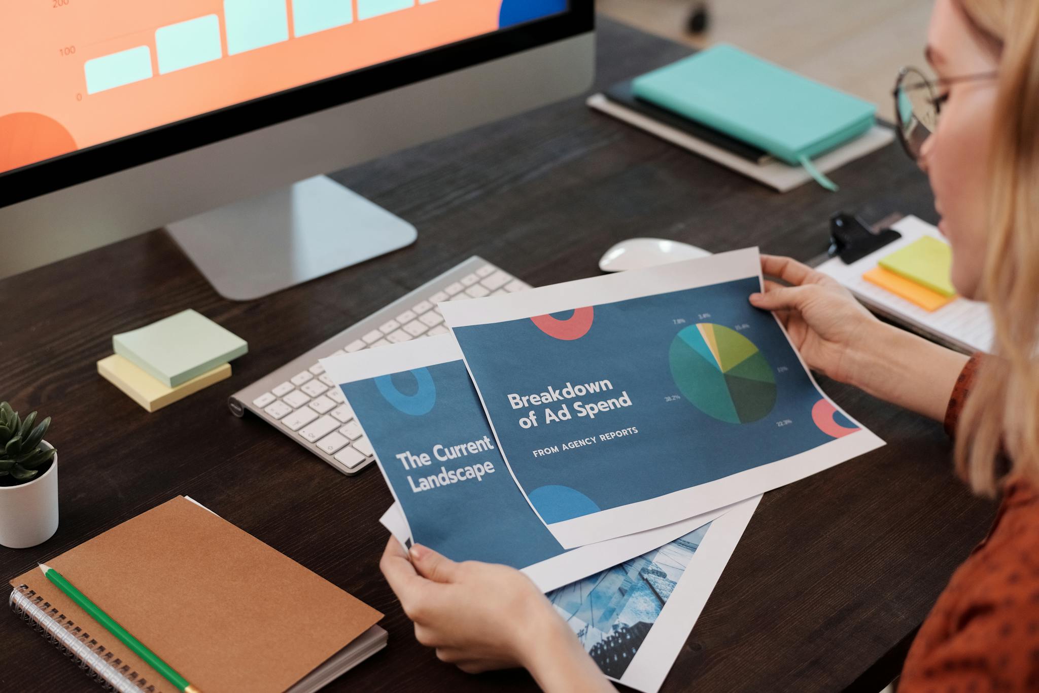 A businesswoman reviews marketing documents with charts at her desk, featuring a computer and office supplies.